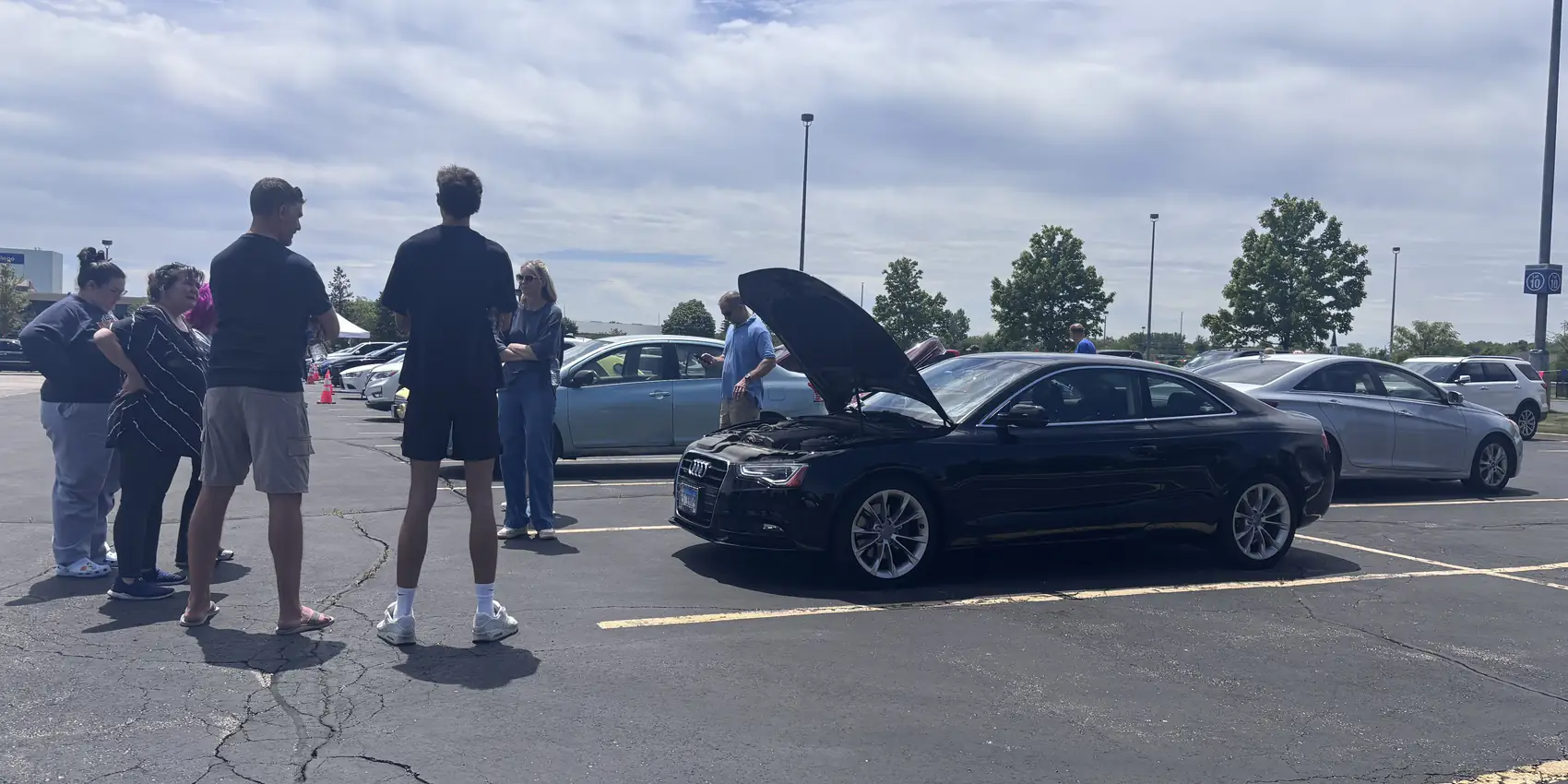 A crowd of people gathered around a black Audi coupe with its hood up at Event #1