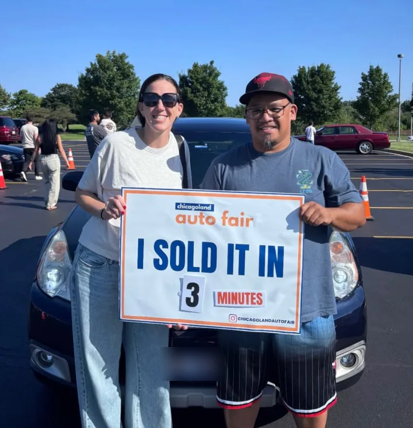 A two-time buyer and the owner she bought from, holding an I sold it in 3 minutes sign in front of the car on the lot at the Auto Fair. Three minutes is the time from parking to the first offer.