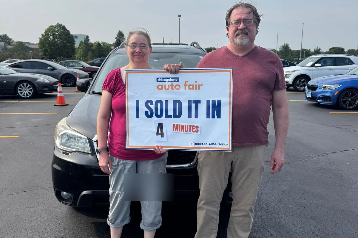Jeffrey and his wife holding a Chicagoland Auto Fair sign reading I sold it in 4 minutes, in front of their black Subaru Forester